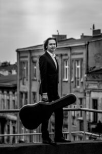 Maciej Staszewski, trzyma w ręku gitarę stoi na balkonie na tle kamienic